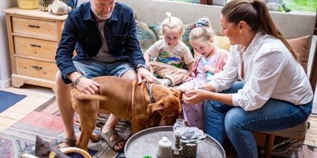 Familie med to børn og to voksne sidder i udestue og kæler med deres hund